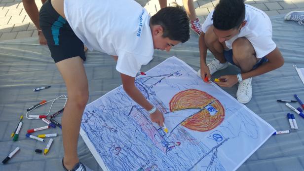 Zwei Jugendliche malen gemeinsam mit Stiften auf einem großen Blatt Papier.