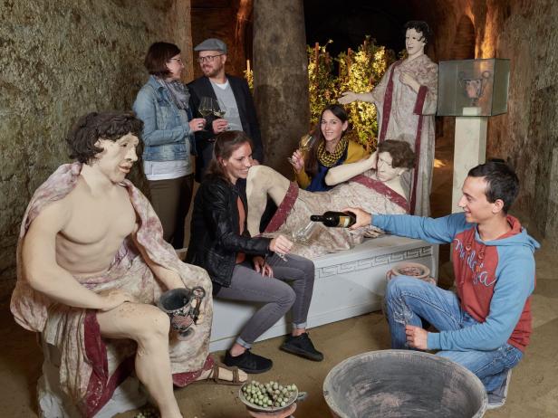 Eine Gruppe von Personen verkostet Wein in einem Keller mit römisch anmutenden Statuen.
