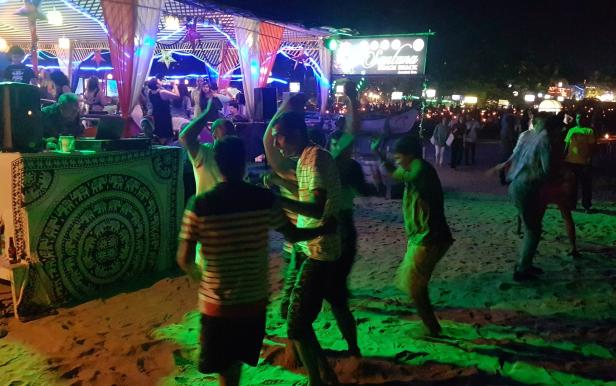 Menschen tanzen nachts am Strand unter bunten Lichtern.