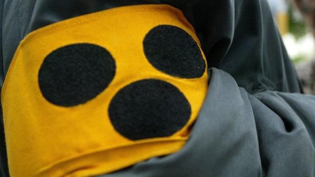 Ein gelbes Stoffabzeichen mit drei schwarzen Punkten ist an einem grauen Kleidungsstück befestigt.