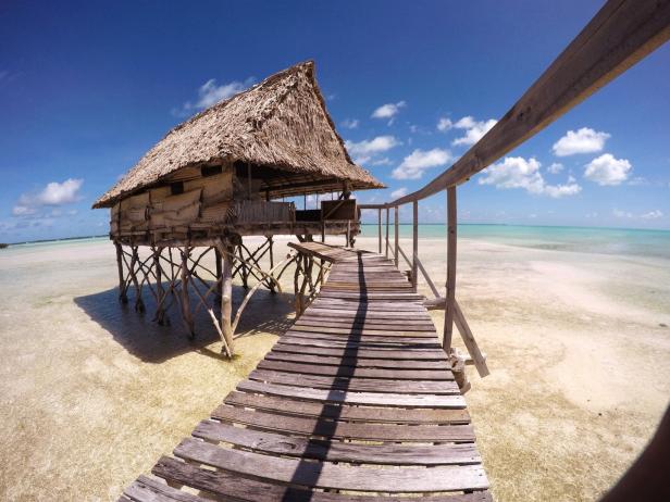 Ein Holzsteg führt zu einem strohgedeckten Haus auf Stelzen im türkisfarbenen Wasser.