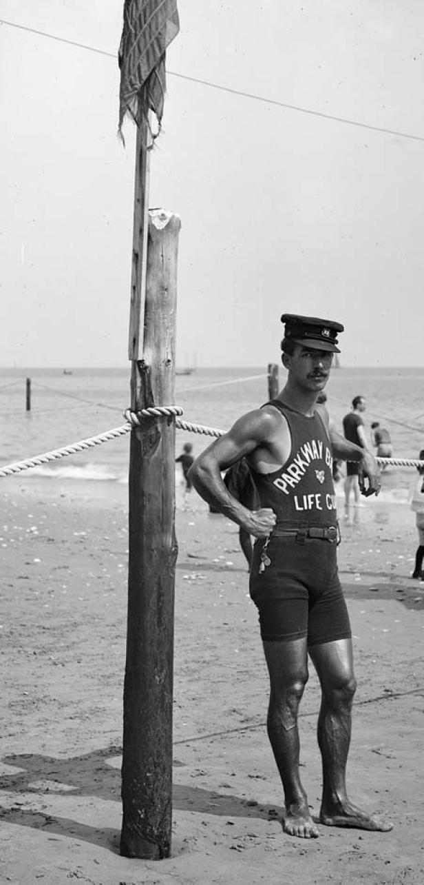 Ein muskulöser Rettungsschwimmer mit Mütze steht an einem Strand.