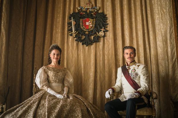 Ein Paar in historischer Kleidung sitzt vor einem goldenen Vorhang mit dem österreichischen Wappen.