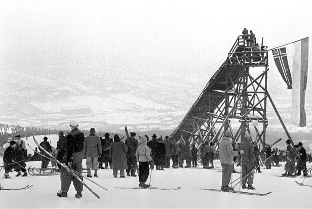 Eine Menschenmenge versammelt sich an einem verschneiten Hang neben einer Skisprungschanze mit isländischer Flagge.