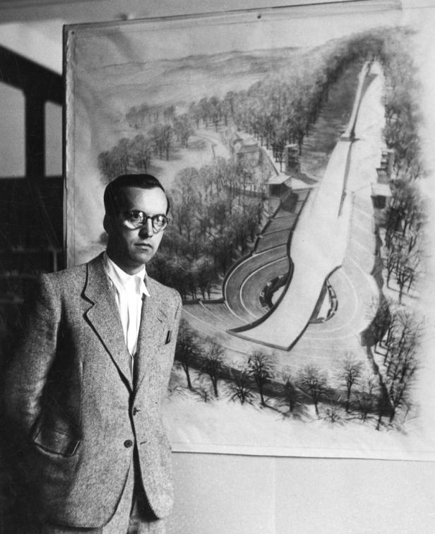 Ein Mann mit Brille steht vor einer Zeichnung eines futuristischen Bauwerks in einer Parklandschaft.