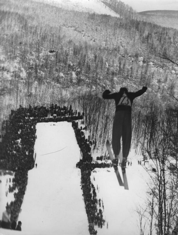 Ein Skispringer in der Luft über einer Zuschauermenge vor einer verschneiten Landschaft.