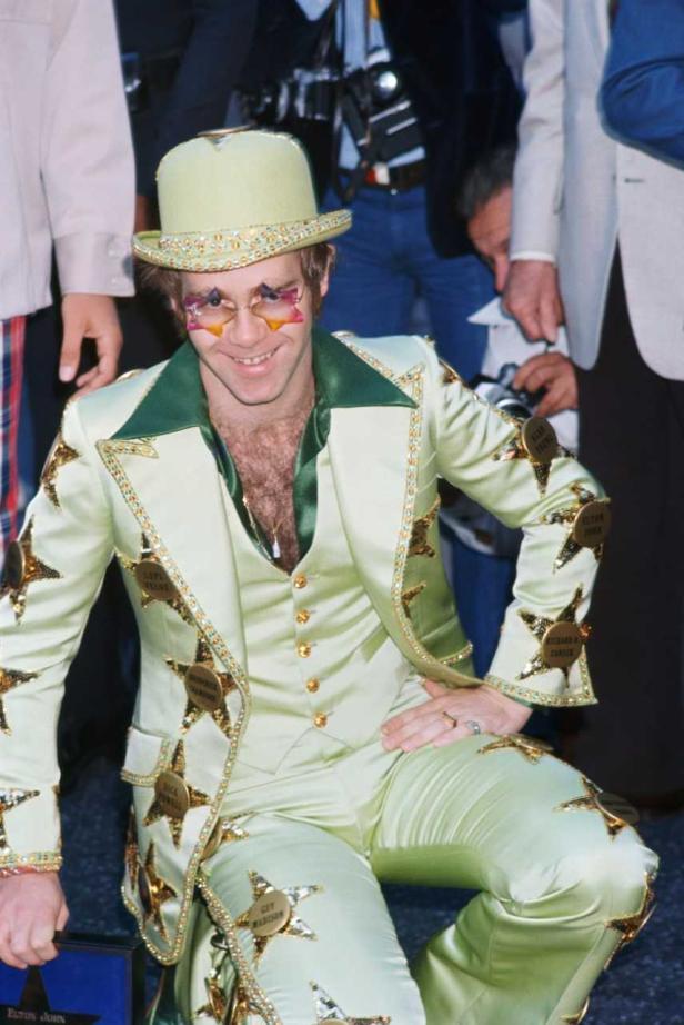 Elton John kniet in einem hellgrünen Anzug mit Sternenapplikationen und trägt eine passende Brille und einen Hut.