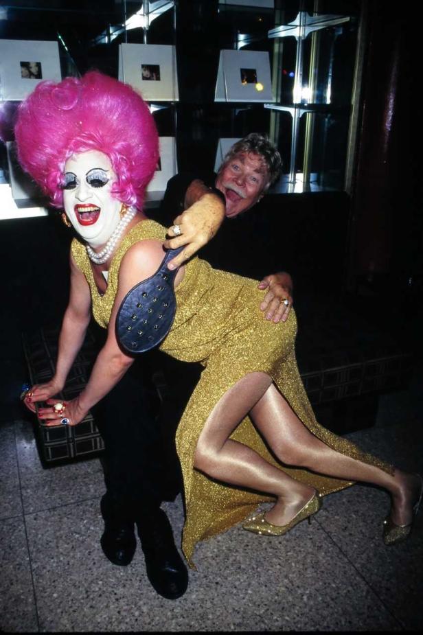 Eine Dragqueen mit pinkfarbener Perücke wird von einem Mann mit einem Paddel gehauen.