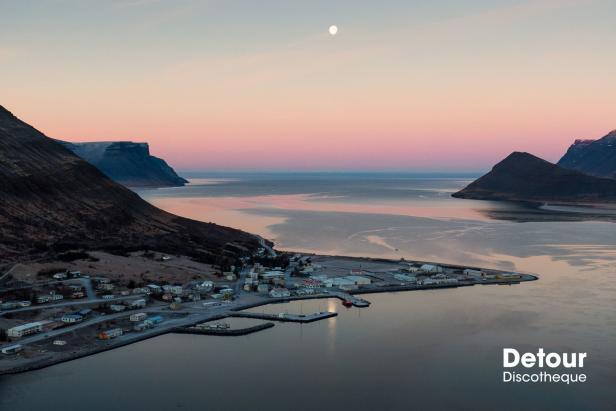 Luftaufnahme einer kleinen Stadt an einer Bucht mit Bergen im Hintergrund bei Sonnenaufgang.