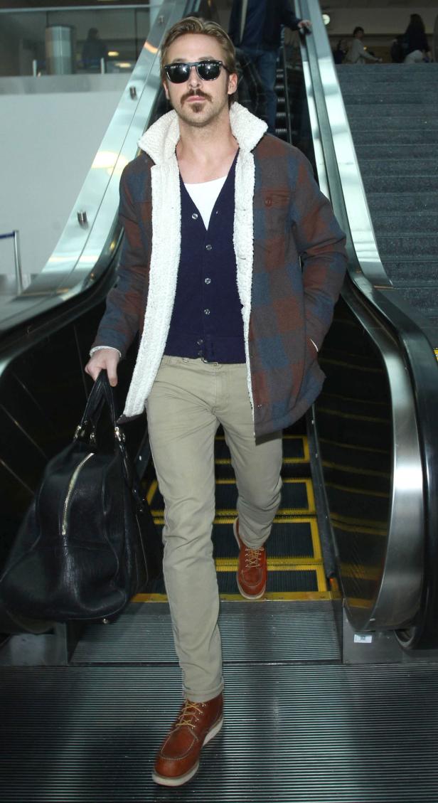 Ryan Gosling mit Sonnenbrille auf einer Rolltreppe mit einer schwarzen Reisetasche.