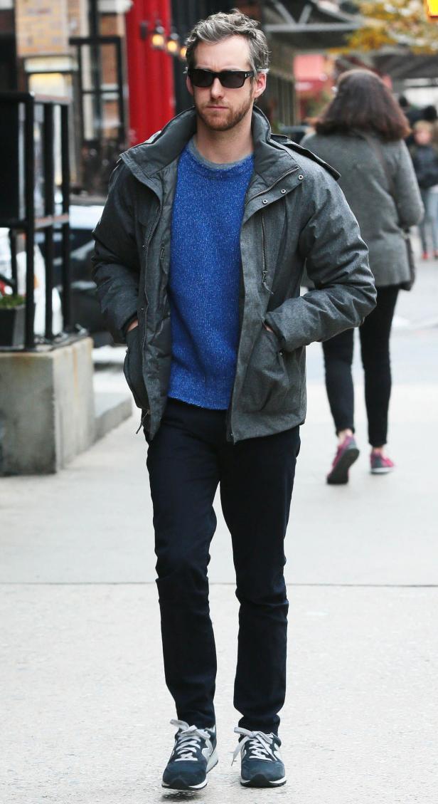 Ein Mann mit Sonnenbrille, blauen Pullover und grauer Jacke geht auf dem Bürgersteig entlang.