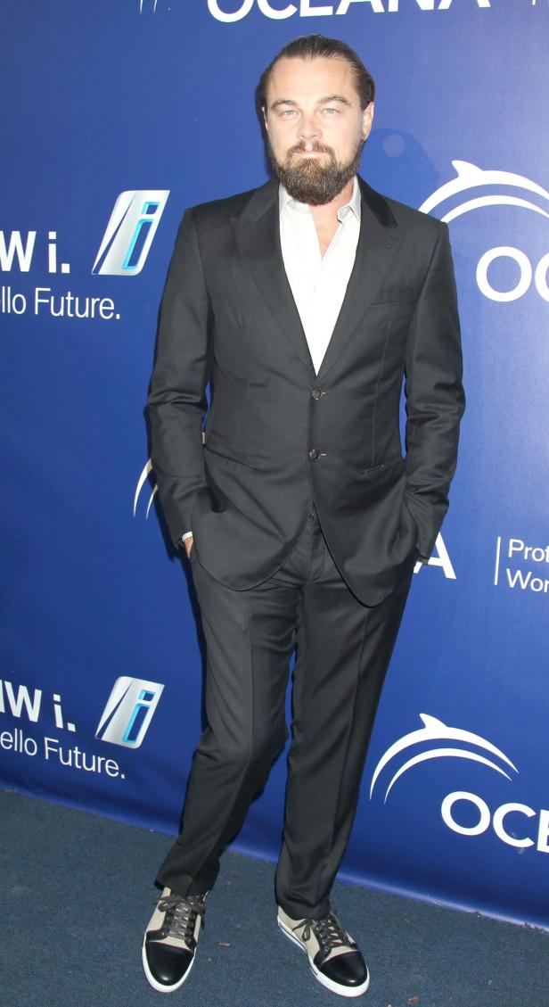 Leonardo DiCaprio in einem dunklen Anzug und Turnschuhen vor einem blauen Hintergrund.