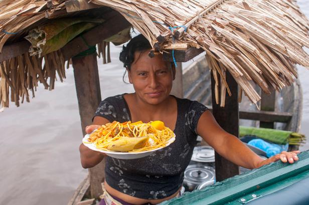 Eine Frau bietet von einem Boot aus ein Teller mit Spaghetti, Kochbanane und Bohnen an.