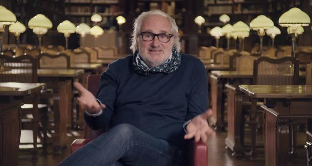 Ein Mann mit Brille sitzt in einer Bibliothek und gestikuliert.