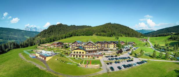 Ein Panoramablick auf ein gelbes Hotelresort in einer grünen Hügellandschaft mit bewaldeten Bergen im Hintergrund.
