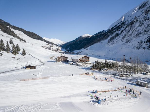 Winterlandschaft mit schneebedeckten Bergen, Häusern und einer Skipiste mit Skifahrern.