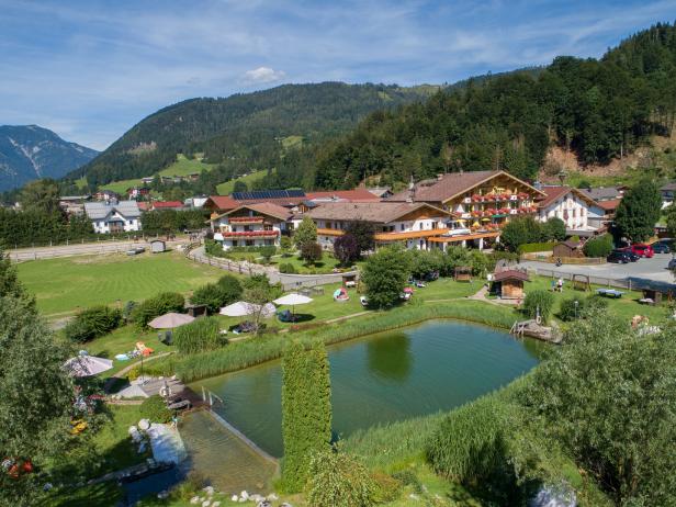 Ein malerischer Ort mit Häusern, einem Teich und grünen Wiesen vor einer bewaldeten Hügellandschaft.