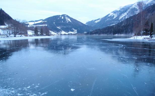 Ein zugefrorener See liegt vor einer verschneiten Berglandschaft.