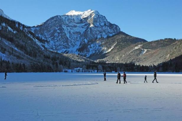 Menschen spazieren auf einem zugefrorenen See vor einer Bergkulisse.