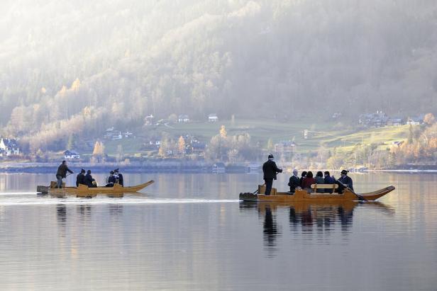 Zwei traditionelle Holzboote fahren auf einem ruhigen See vor einer bewaldeten Hügellandschaft.
