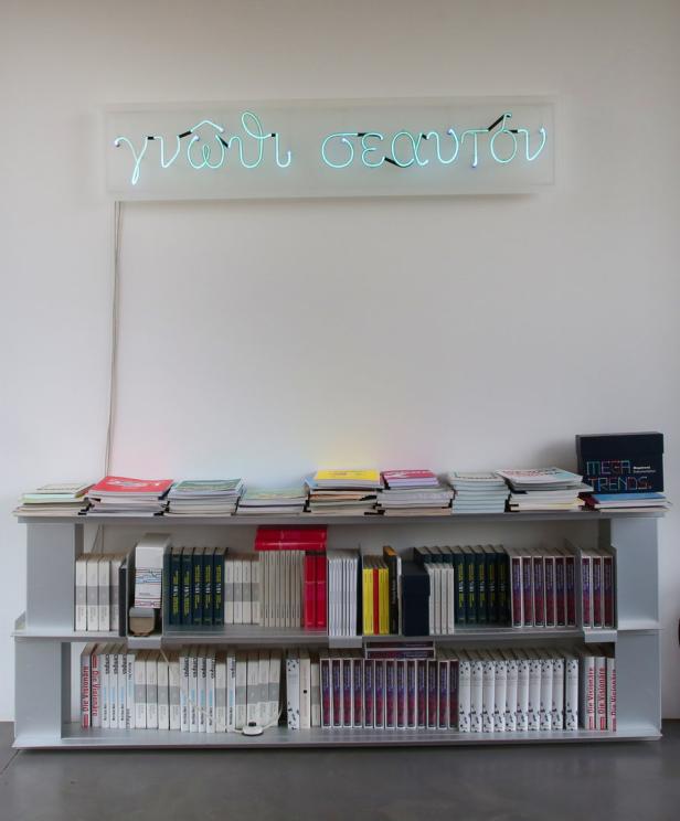 Ein Bücherregal unter einer Neonreklame mit der griechischen Aufschrift „γνῶθι σεαυτόν“ (Erkenne dich selbst).