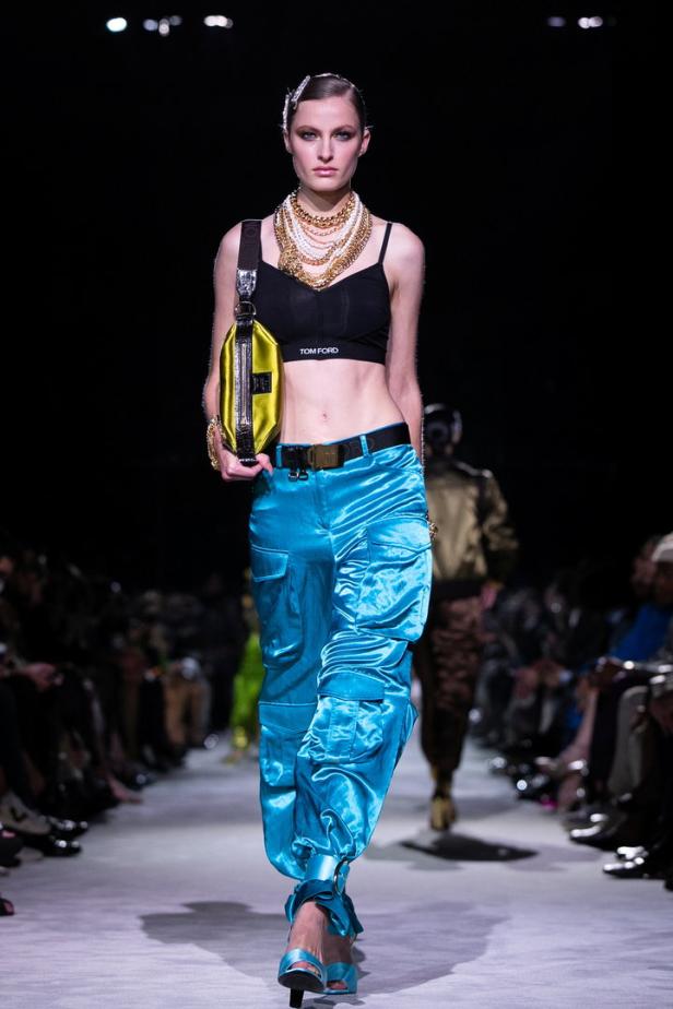 Ein Model auf dem Laufsteg trägt ein Outfit von Tom Ford mit türkisfarbener Hose und goldenen Accessoires.