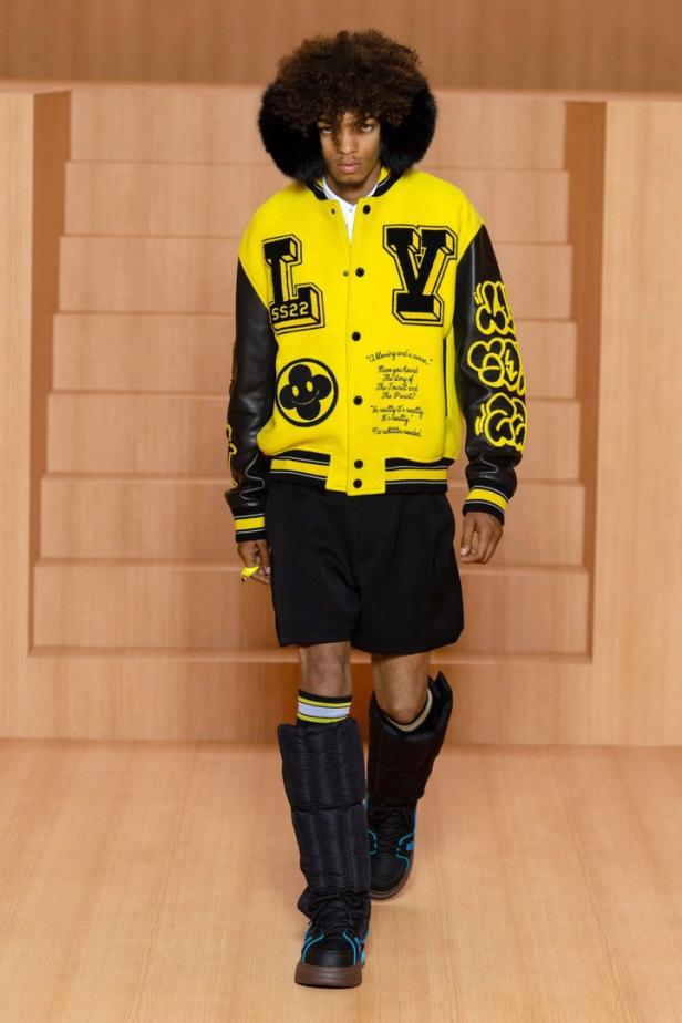 Ein Mann in einer gelben Louis Vuitton Jacke mit schwarzen Shorts und Stiefeln auf dem Laufsteg.