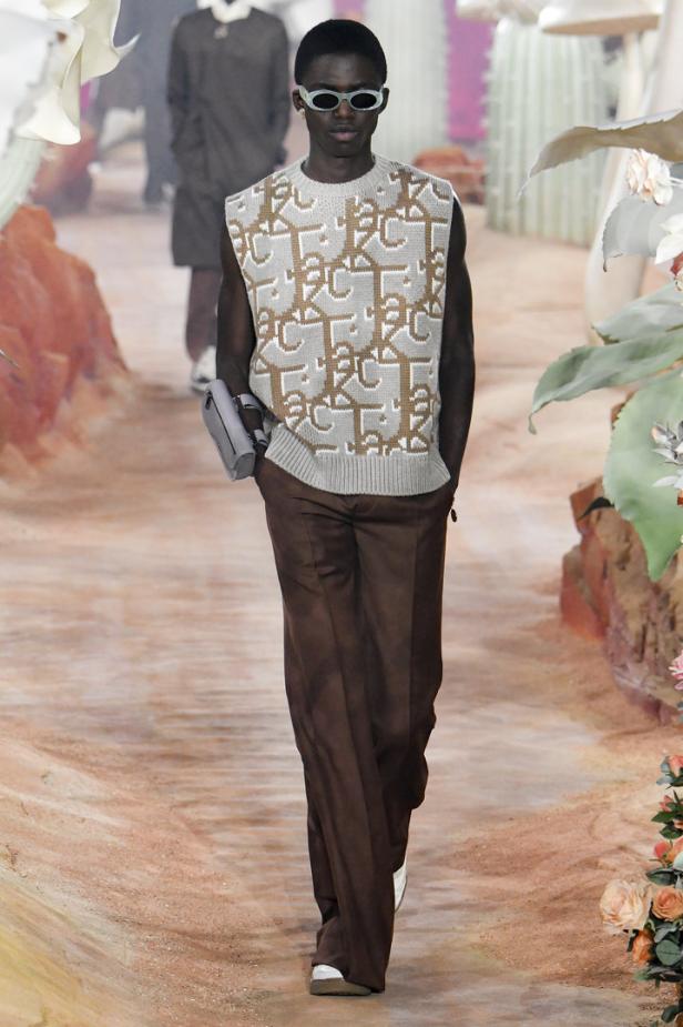 Ein Mann mit Sonnenbrille und ärmellosem Pullover mit „Dior“-Muster läuft auf einem Laufsteg.