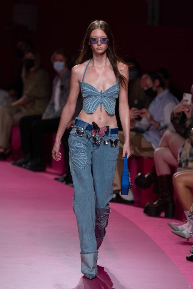Ein Model präsentiert ein Jeans-Ensemble mit Schmetterlingsmotiven auf einem pinkfarbenen Laufsteg.