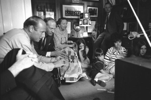 Eine Gruppe von Menschen, darunter Kinder, schaut aufmerksam auf einen Fernseher.