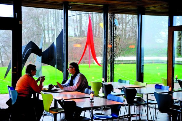 Zwei Personen sitzen in einem Café mit Blick auf eine grüne Wiese mit Kunstobjekten.