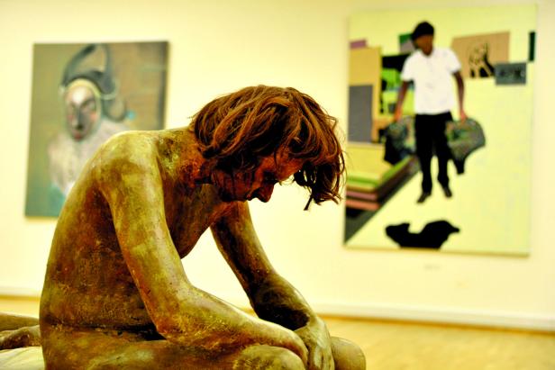 Eine bronzefarbene Skulptur einer sitzenden, nachdenklichen Person vor zwei Gemälden.