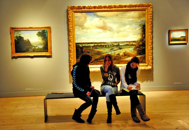 Drei junge Frauen sitzen auf einer Bank vor Gemälden in einem Museum.