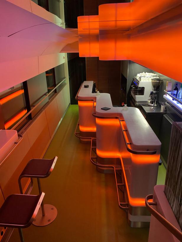 Eine Bar mit beleuchteten Tresen und Hockern in einem futuristischen Design.