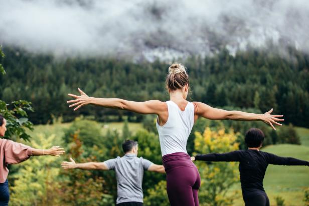 Eine Gruppe von Menschen praktiziert Yoga im Freien vor einer bewaldeten Bergkulisse.