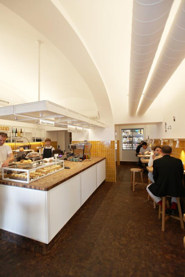 In einem Café bedient ein Mann Kunden an der Theke, während andere Gäste an Tischen sitzen.
