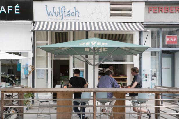 Drei Personen sitzen an einem Tisch vor dem Restaurant „Wulfisch“.