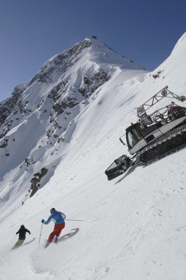 Zwei Skifahrer fahren einen steilen Hang neben einer Pistenraupe hinunter.