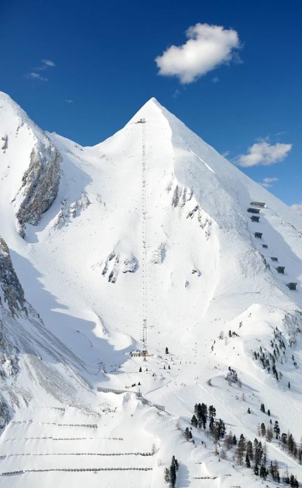 Eine lange Seilbahn führt auf einen schneebedeckten Berggipfel unter blauem Himmel.
