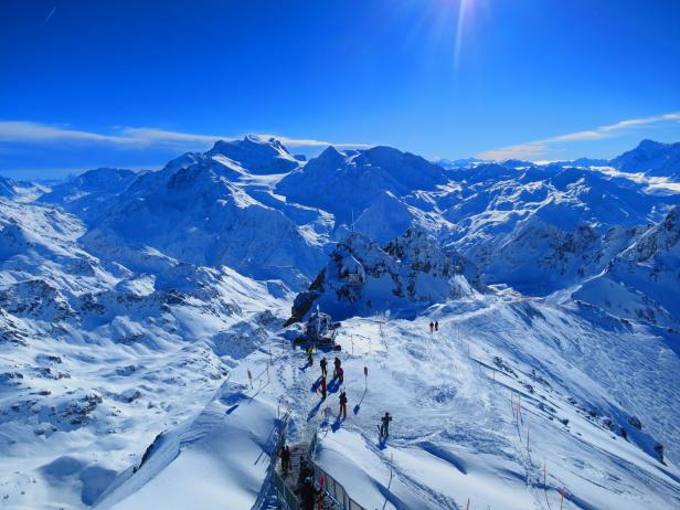 Blick von einem schneebedeckten Berggipfel auf eine weite Berglandschaft unter blauem Himmel.
