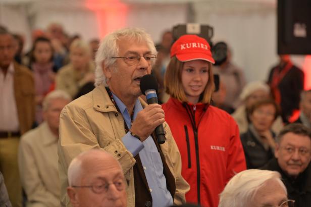 Ein Mann spricht in ein Mikrofon, während eine junge Frau in roter Jacke daneben steht.