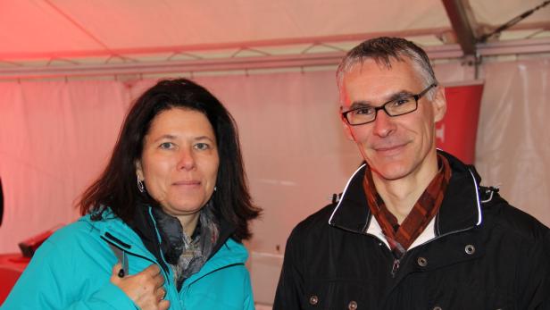 Eine Frau in einer türkisfarbenen Jacke und ein Mann mit Brille stehen nebeneinander.