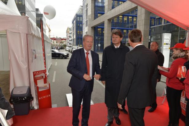 Männer in Anzügen betreten einen roten Teppich vor einem Gebäude.