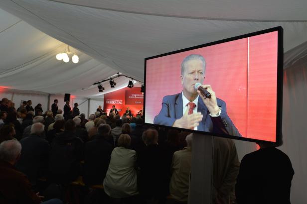 Ein Mann spricht auf einem Bildschirm vor Publikum in einem Zelt.