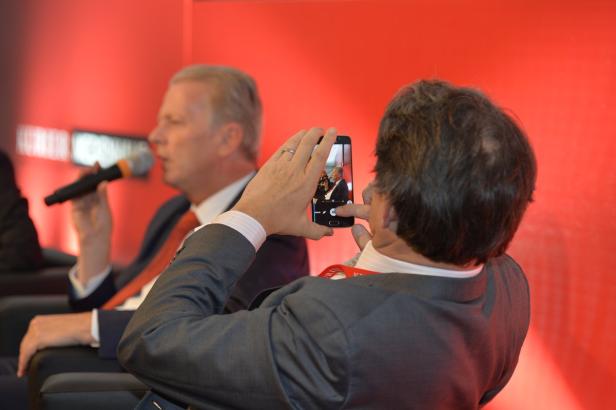 Ein Mann fotografiert mit seinem Smartphone einen Redner auf einer Bühne.