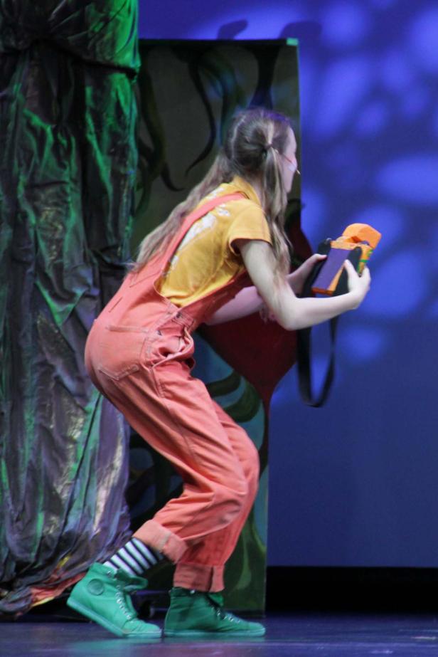 Eine junge Frau in orangefarbener Latzhose hält eine Requisite auf einer Theaterbühne.