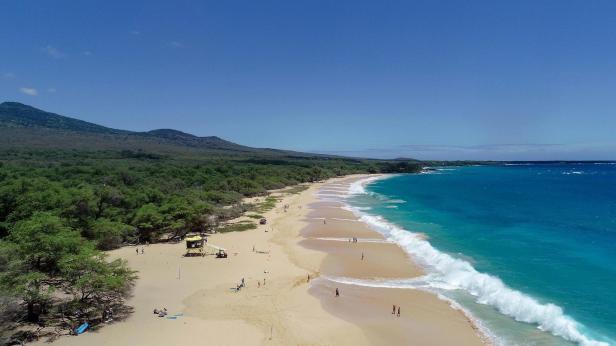 Ein Sandstrand mit türkisfarbenem Wasser und einem bewaldeten Hügel im Hintergrund.