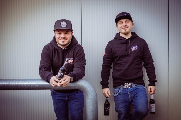 Zwei Männer posieren mit „Keck Kola“ vor einer grauen Wand.