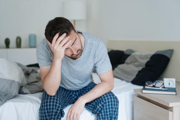Ein Mann sitzt mit dem Kopf in der Hand auf dem Bett und wirkt verzweifelt.