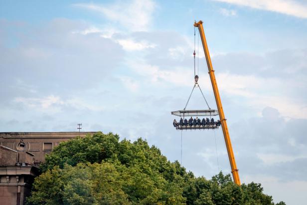 Ein Kran hebt eine Plattform mit Menschen in die Luft, über Bäumen und einem Gebäude.